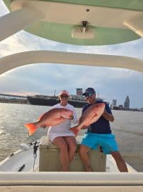 Two anglers proudly displaying their Red Snapper catch while enjoying a fishing trip on a boat in a scenic urban backdrop.