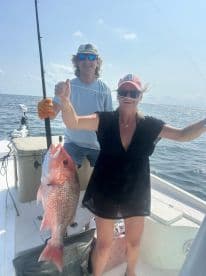 A woman proudly displays a large Red Snapper while fishing on a boat, with a scenic ocean backdrop.