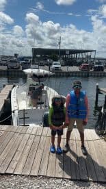 Two young anglers in life jackets pose on the dock next to a fishing boat, ready for a day on the water.