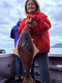An angler proudly displaying a large Halibut while fishing on a boat in a scenic coastal environment.