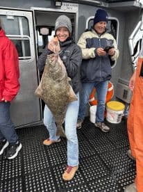 An angler proudly displaying a large Halibut on a fishing charter, surrounded by fellow anglers on the deck.