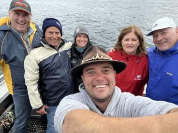 Group of six smiling individuals posing for a selfie on a fishing boat, enjoying a day out on the water.