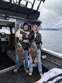 Two anglers proudly displaying their catch of Halibut while aboard a fishing boat in a scenic coastal environment.