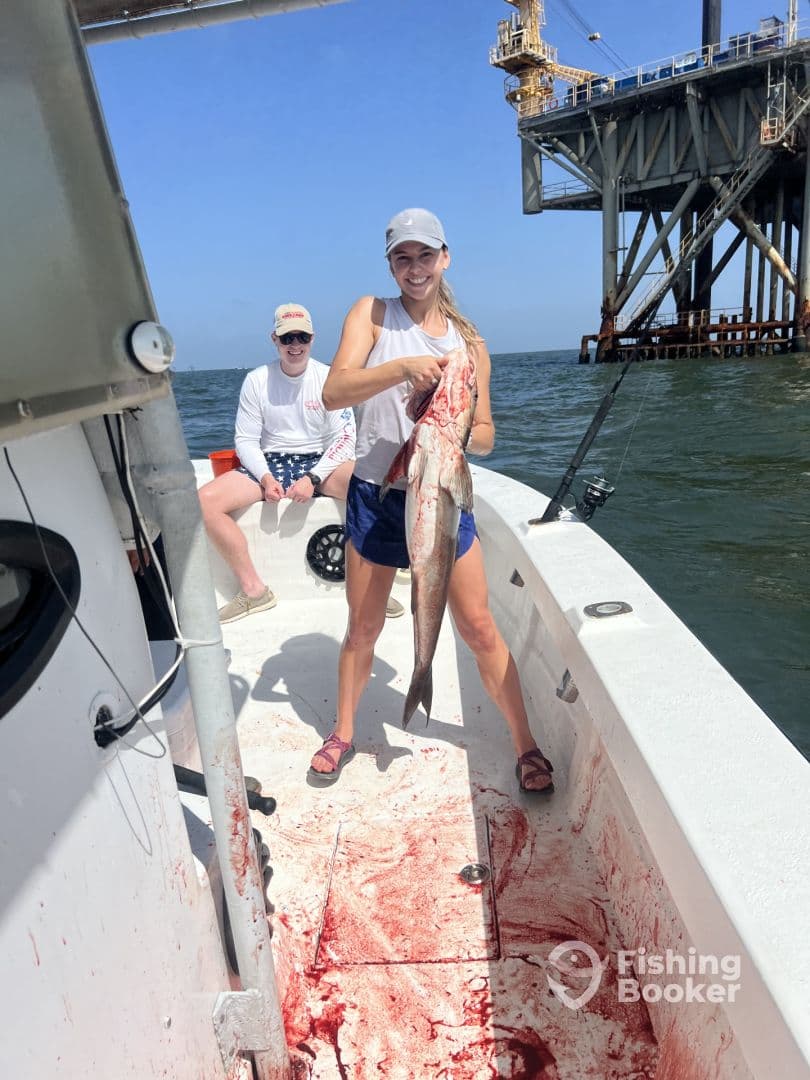 A smiling angler proudly displays a large fish on a fishing boat, with an oil rig in the background, showcasing a successful day on the water.