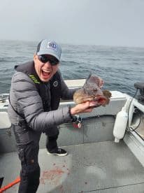 Excited angler holding a large Lingcod aboard a fishing boat in foggy waters, showcasing a successful catch.