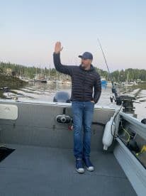 A man stands on the interior of a fishing boat, waving, with a scenic harbor filled with boats in the background.