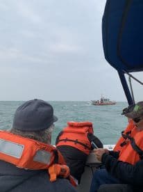 Passengers wearing life jackets aboard a fishing boat, observing another vessel in the distance on a cloudy day.