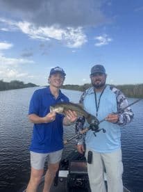 Two anglers proudly displaying a large Largemouth Bass while fishing in a scenic waterway.