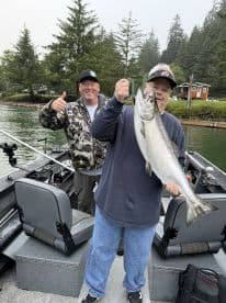 Two anglers celebrating a successful catch of a large Salmon while fishing from a boat in a scenic coastal environment.