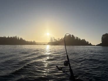 A serene sunset fishing scene with a fishing rod bent, indicating a catch in progress, surrounded by a picturesque coastal landscape.