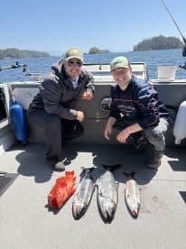 Two anglers proudly displaying their catch of three Salmon and a Rockfish aboard a fishing boat in a scenic coastal environment.