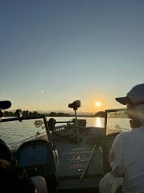 Two anglers enjoying a sunset view from inside a fishing boat, with the serene water and mountains in the background.