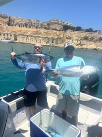 Two anglers proudly displaying their catches of large tuna while on a boat, with a scenic coastal backdrop.