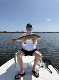 An angler proudly displaying a large Redfish while seated on a boat in a scenic fishing environment.