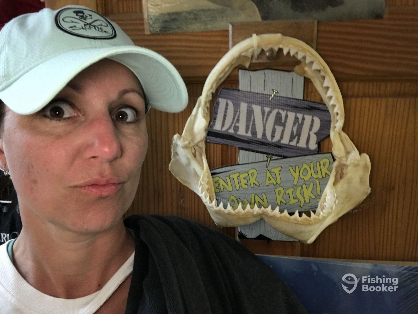 A woman poses with a shark jaw and a humorous warning sign in a fishing-themed setting.