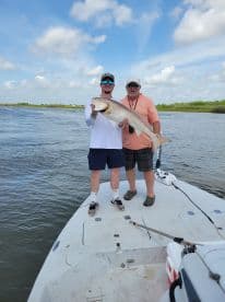 Two anglers proudly displaying a large Redfish on the bow of a boat during a fishing trip.