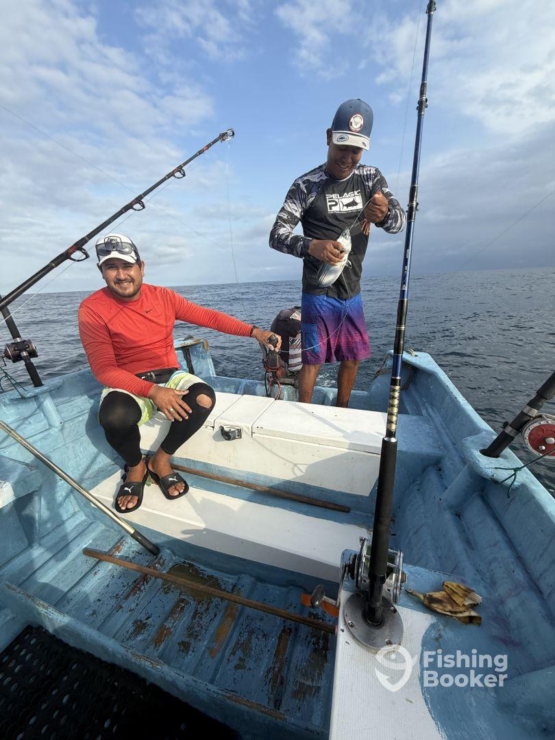 Two anglers aboard a small fishing boat, one preparing a catch while the other relaxes. The scene captures the camaraderie of fishing in open waters.