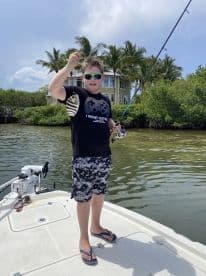 A young angler proudly displaying a caught fish while fishing from a boat in a tropical setting, surrounded by lush greenery.
