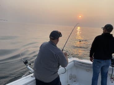 Two anglers actively fishing at sunset, with one reeling in a catch while the other observes from the boat.