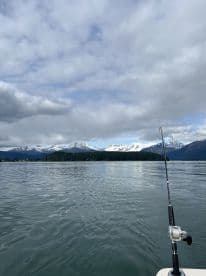 Scenic view of a fishing environment with mountains and cloudy skies, featuring a fishing rod positioned on the boat.