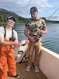 Two anglers on a boat displaying a freshly caught fish, surrounded by a scenic backdrop of mountains and water.
