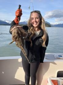 A smiling angler proudly displays a large Halibut while fishing in a scenic Alaskan backdrop.