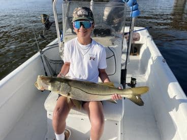 A young angler proudly displaying a large Snook while seated on a boat, showcasing a successful day of fishing.