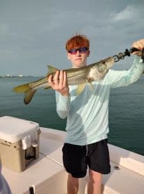 A young angler proudly displaying a Snook caught while fishing from a boat, showcasing a beautiful coastal backdrop.