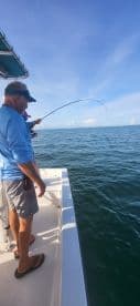 Two anglers engaged in fishing action on a boat, with one reeling in a catch against a backdrop of clear blue skies and calm waters.