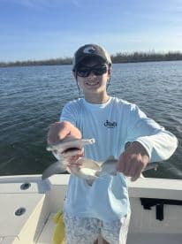 A young angler proudly displaying two caught sharks while on a fishing trip, showcasing the excitement of fishing in a coastal environment.