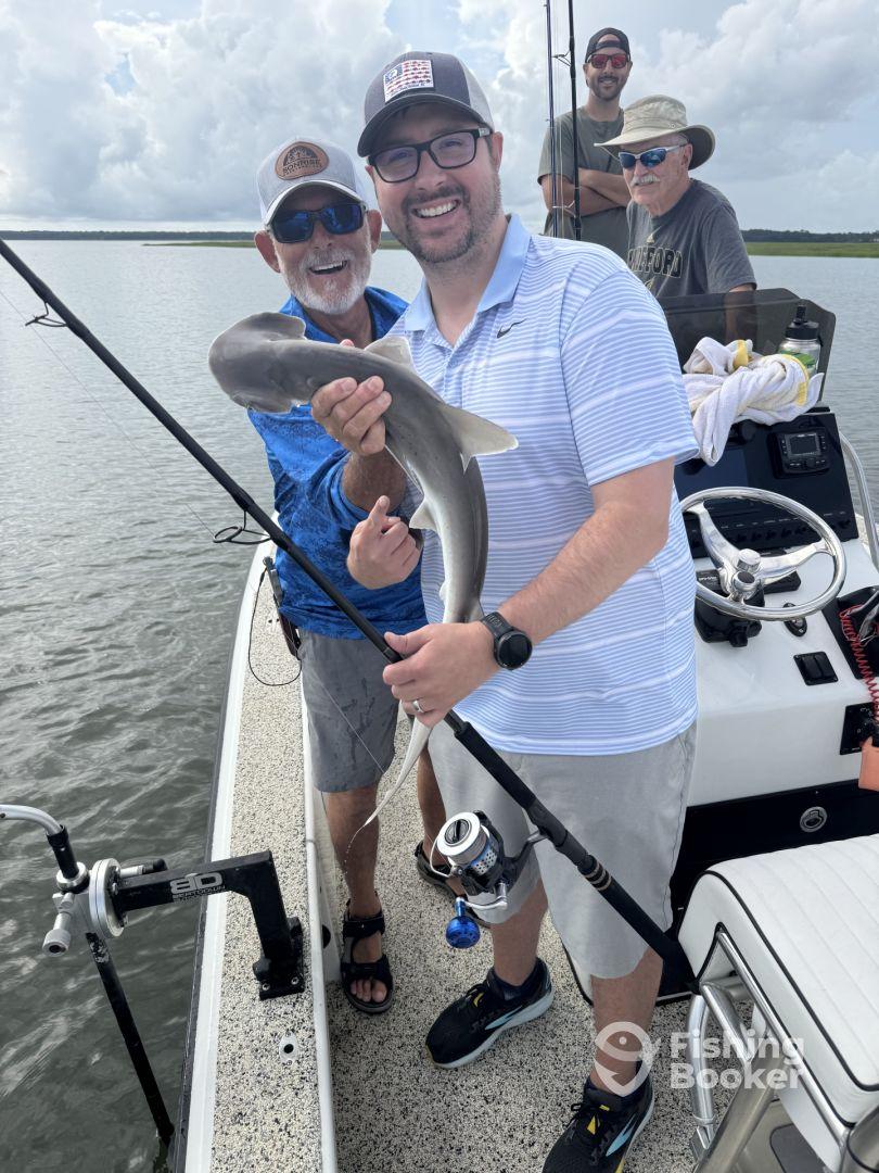 Two anglers proudly displaying a caught shark while fishing on a boat in a scenic waterway.