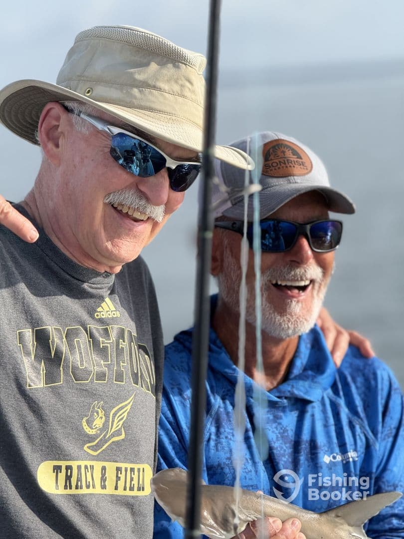 Two anglers smiling while holding a small shark on a fishing trip, showcasing a successful catch in a sunny marine environment.