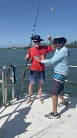 Two anglers enjoying a lively moment on the boat, one proudly displaying a catch while the other assists, set against a scenic water backdrop.
