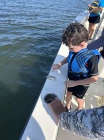 A young boy in a life jacket is engaged in fishing, pointing at a small fish on the boat's edge during a family fishing trip.