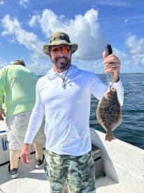 An angler proudly displaying a Flounder while fishing on a sunny day, with a scenic backdrop of blue skies and water.