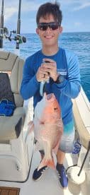 A young angler proudly displaying a large Snapper while fishing on a boat in clear blue waters.