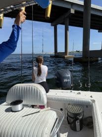 A young angler and an adult are fishing under a bridge, with fishing rods set up in a boat, showcasing a typical day of fishing in a coastal environment.