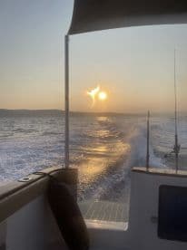 A scenic view from the side of a boat during sunset, showcasing the tranquil waters and the sun reflecting off the waves.