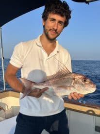 An angler proudly displaying a Snapper caught during a fishing trip on a boat, with a clear blue ocean backdrop.