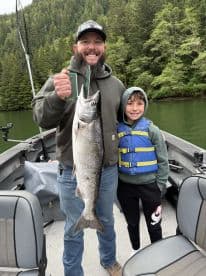 A proud father and son duo displaying a large Salmon caught during their fishing trip in a scenic river setting.