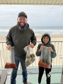 A father and son duo proudly displaying their catch of crabs in bags, with a scenic beach backdrop after a successful fishing trip.