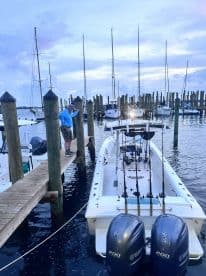 A fisherman preparing to board a center console boat at a marina during twilight, surrounded by sailboats and fishing vessels.