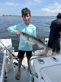 A young angler proudly displaying a large Striped Bass while fishing from a boat, showcasing a sunny day on the water.