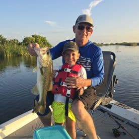 A young boy and his grandfather proudly display a large Largemouth Bass while fishing on a serene lake, showcasing a memorable family fishing experience.