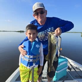 A young boy in a life jacket proudly poses with his grandfather, showcasing a large Largemouth Bass caught during a serene day on the water.