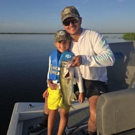A young angler and an adult proudly displaying a large Largemouth Bass while fishing on a calm lake.
