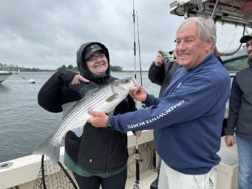 A happy angler proudly displaying a striped bass while fishing on a cloudy day, surrounded by fellow anglers on a boat.