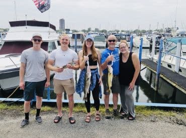 A group of four adults proudly displaying their catch of the day, including a large Walleye, at the marina after a successful fishing trip.