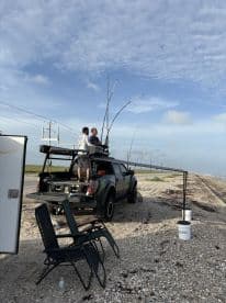 Two anglers fishing from the top of a truck with long rods, enjoying a day by the shore.