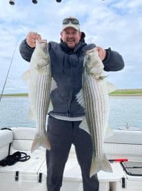 Angler proudly displaying two large Striped Bass while aboard a fishing boat, showcasing a successful day on the water.